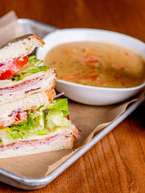 Stacked deli sandwich with lettuce, tomato, and sliced meats, served with a bowl of soup and iced drink from Solare Café at Element Eatery food hall
