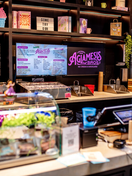 Agla-Mesis Bros ice cream and chocolate counter at Element Eatery food hall, featuring colorful signage, digital menu boards, display case with sweets, and a freezer stocked with ice cream pints