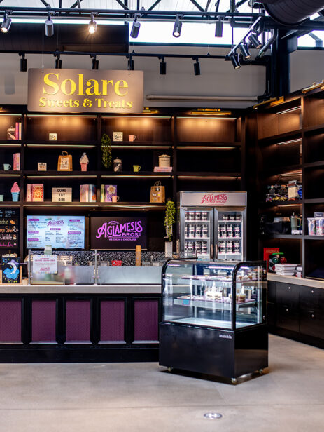 Agla-Mesis Bros ice cream and chocolate counter at Element Eatery food hall, featuring colorful signage, digital menu boards, display case with sweets, and a freezer stocked with ice cream pints