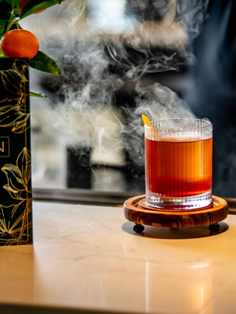 Smoked craft cocktail garnished with an orange peel, served on a wooden coaster next to The Garden’s floral menu at Element Eatery food hall
